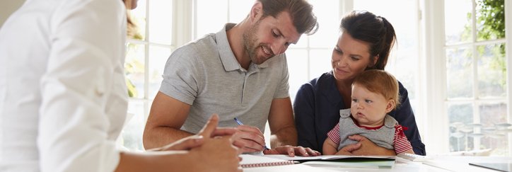 A man sitting next to his wife and baby at a sunny kitchen counter and signing documents from a female mortgage lender.