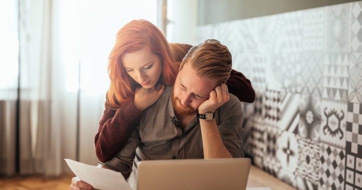 A woman leaning over a man's shoulder as he sits in front of a laptop with his head resting in his hand and holds up a piece of paper.