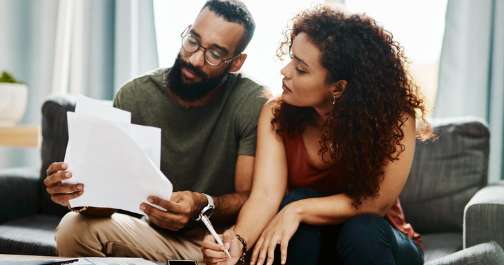A man and woman sitting on their couch looking through bills and signing papers.