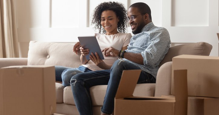 A couple looks at a pad while sitting on a couch near moving boxes