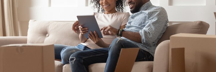 A couple looks at a pad while sitting on a couch near moving boxes