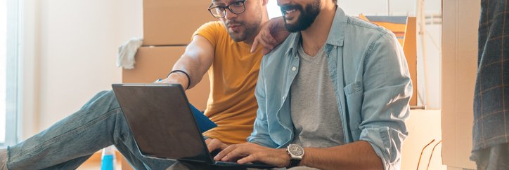 Two people sitting on the floor surrounded by moving boxes while looking up something on a laptop.