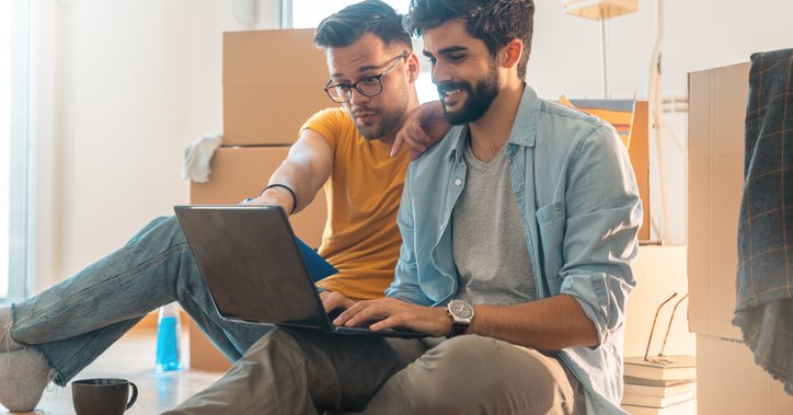 Two people sitting on the floor surrounded by moving boxes while looking up something on a laptop.