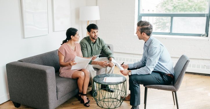 A man and woman sitting on a couch and talking with their realtor while holding paperwork.