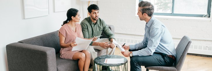 A man and woman sitting on a couch and talking with their realtor while holding paperwork.