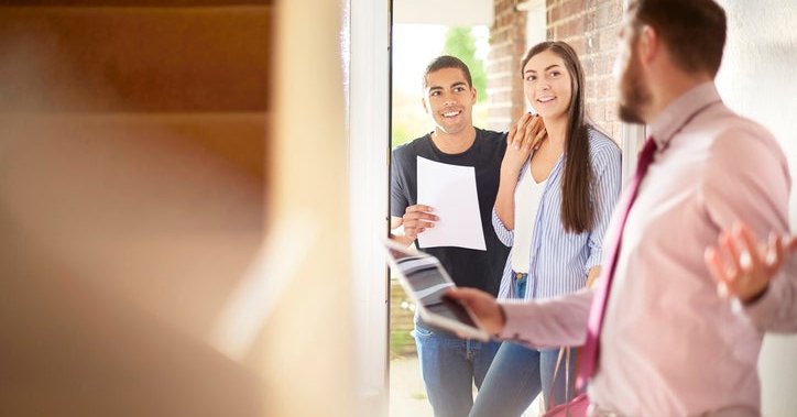 A man and woman looking through the door of a house held open by a realtor.