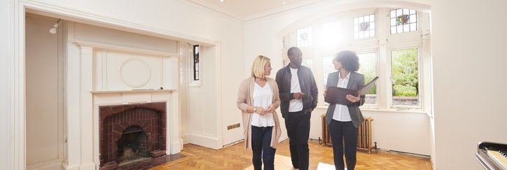 A couple speaking with their realtor while looking around the empty living room of a home.