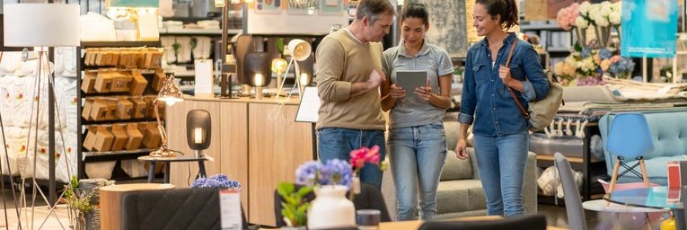 A man and woman walking around a furniture store and looking at a tablet held out by a saleswoman.