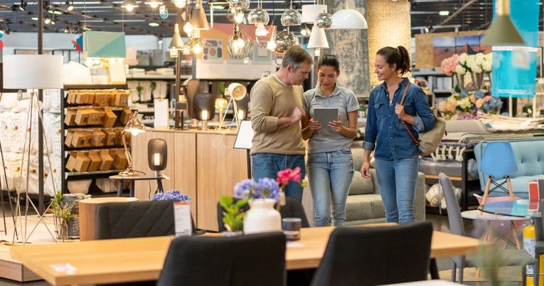 A man and woman walking around a furniture store and looking at a tablet held out by a saleswoman.