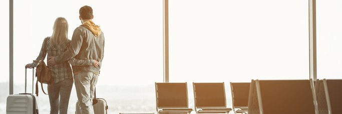 A man and woman standing with their arms around each other and their suitcases next to them at a large airport window.