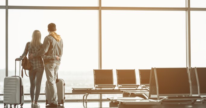 A man and woman standing with their arms around each other and their suitcases next to them at a large airport window.