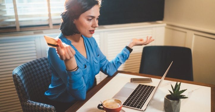 A woman looking frustrated with a credit card in her hand while looking at a laptop.