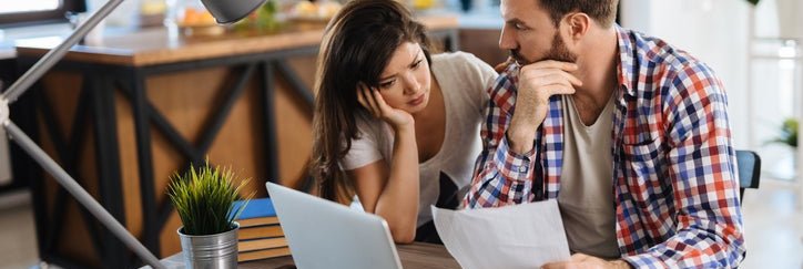A worried couple looks at bills, with a laptop and a credit card on the table.