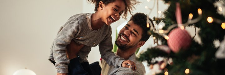 A smiling dad holding up his laughing daughter while they decorate a Christmas tree.