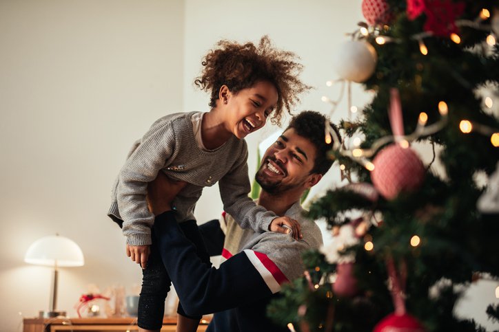 A smiling dad holding up his laughing daughter while they decorate a Christmas tree.