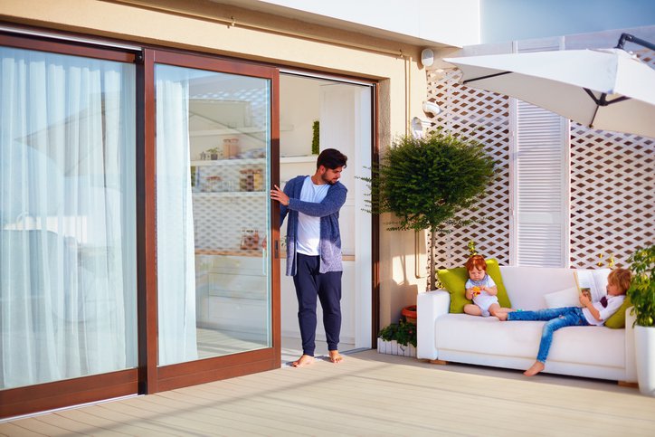 A dad opening a glass sliding door to check on his two young children sitting on a couch on the back deck.