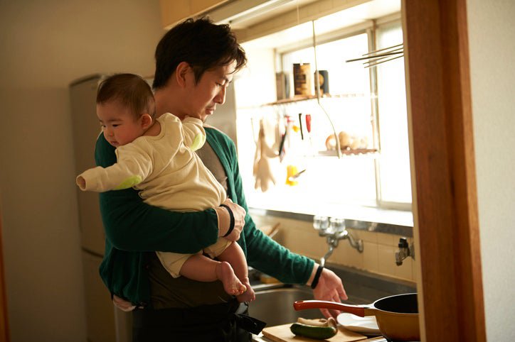 A parent holding a baby in one arm while cooking in a small kitchen.