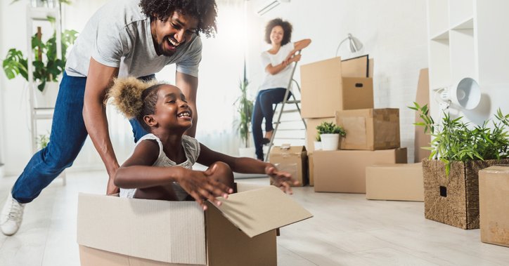 A dad pushing his laughing daughter along the floor in a moving box while the mom looks on smiling while packing other boxes.