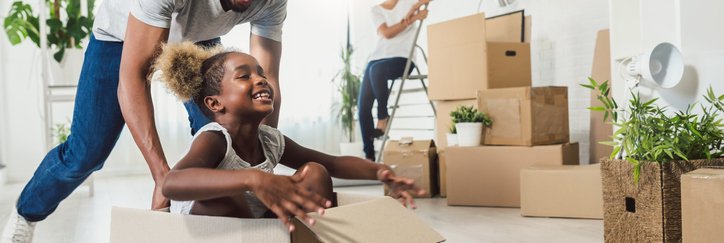A dad pushing his laughing daughter along the floor in a moving box while the mom looks on smiling while packing other boxes.