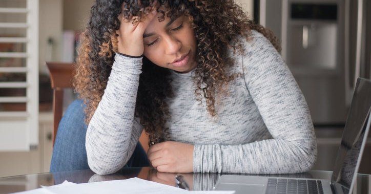 A woman looking worried, at a table with papers and a laptop.