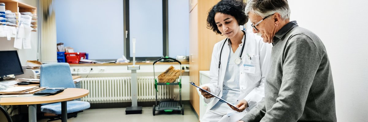 A doctor sitting next to a patient in an exam room and going over test results on a clipboard.