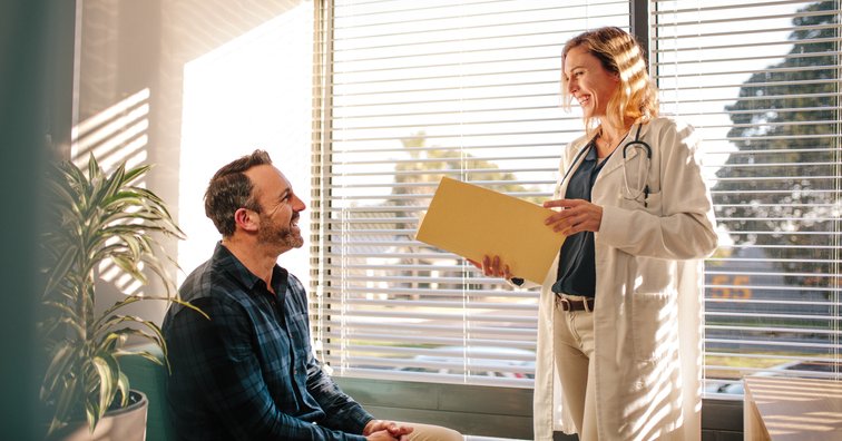 A doctor standing in front of a sunny window in her office and speaking with her male patient sitting in a chair.