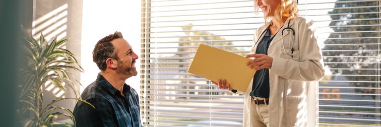 A doctor standing in front of a sunny window in her office and speaking with her male patient sitting in a chair.