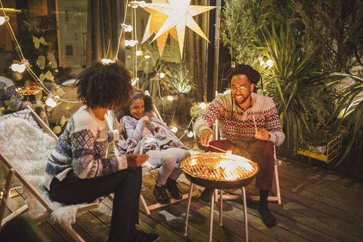 Two parents and their child making smores on their backyard patio decorated with twinkle lights.