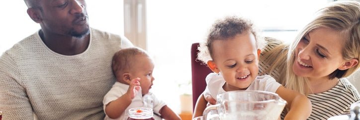 A mom and dad smiling and each holding their toddler and baby while eating breakfast at a sunny table.