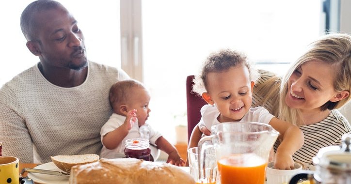 A mom and dad smiling and each holding their toddler and baby while eating breakfast at a sunny table.