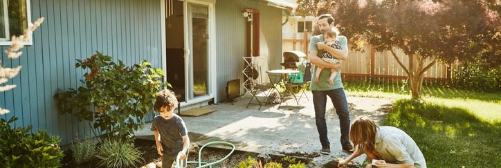 A mom working in the backyard garden with a toddler while the dad walks out of the house holding a baby.