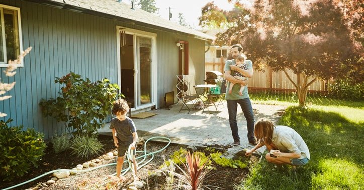 A mom working in the backyard garden with a toddler while the dad walks out of the house holding a baby.