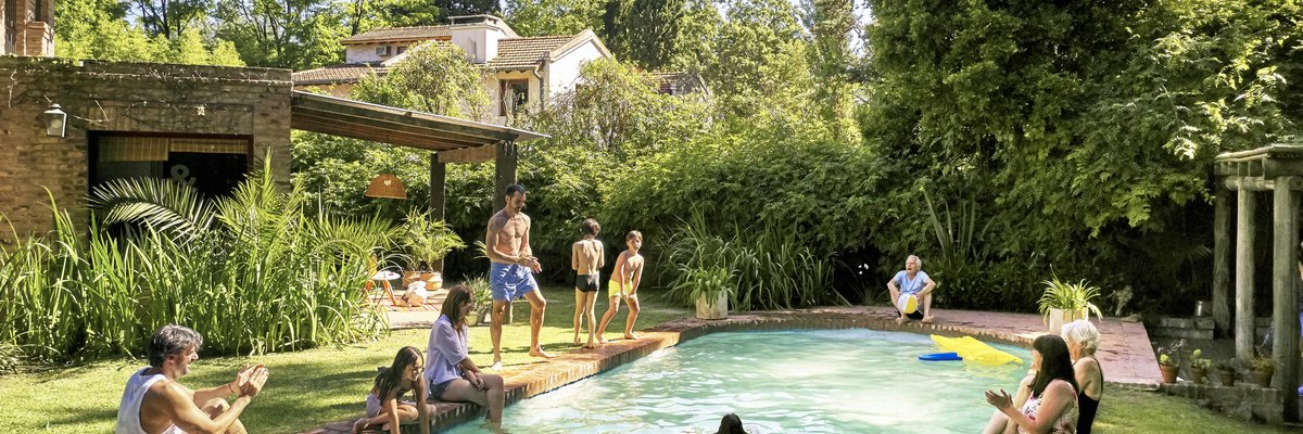 A family playing in a backyard pool surrounded by green bushes and trees.
