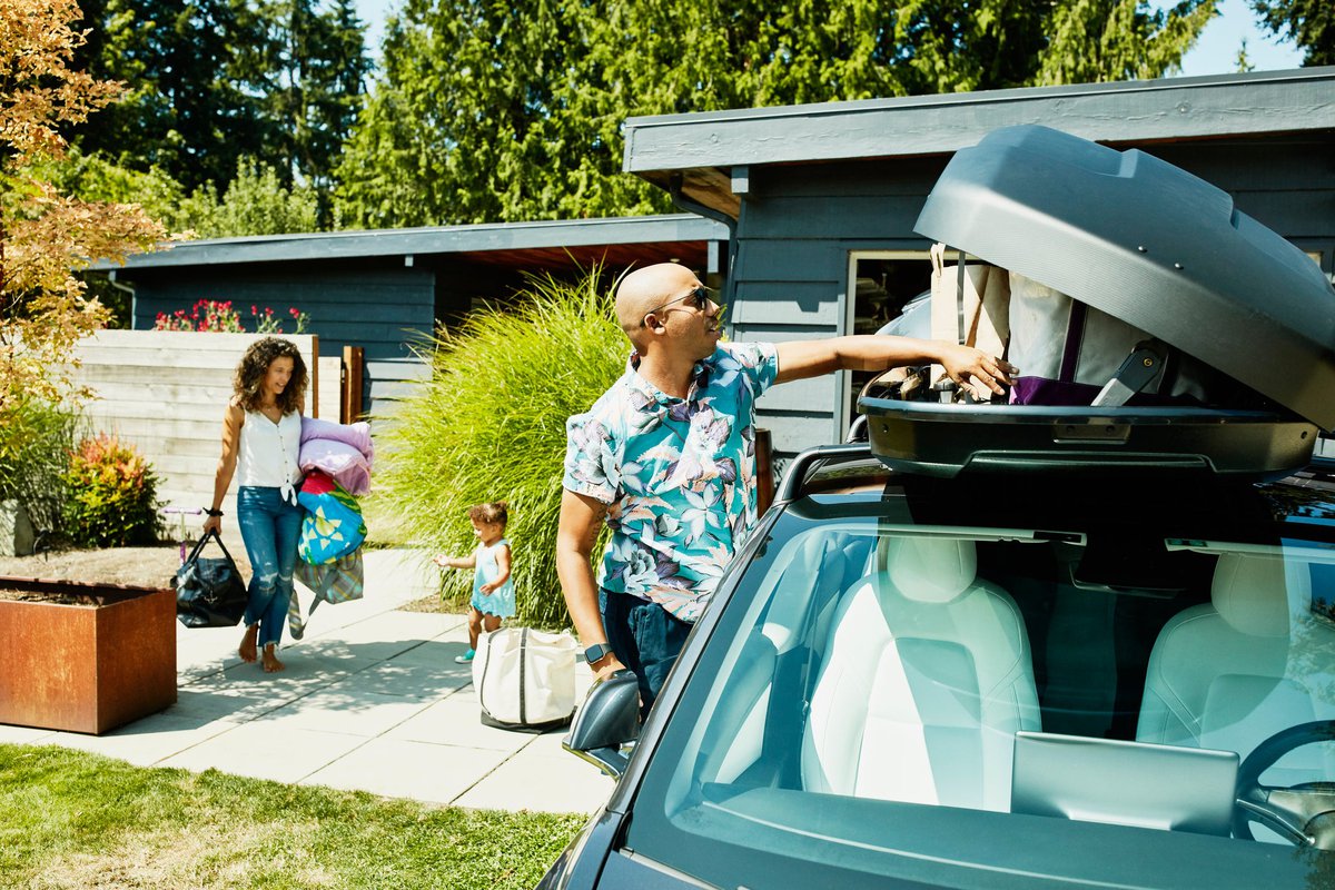 A family loads bags into a car in its driveway, preparing for a road trip.
