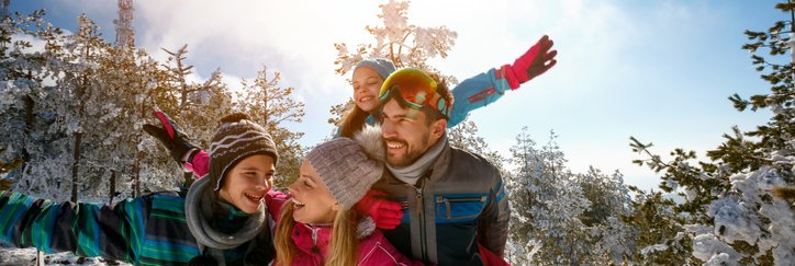A mom, dad, son, and daughter taking a break from downhill skiing to play in the snow on a sunny mountain slope surrounded by trees.
