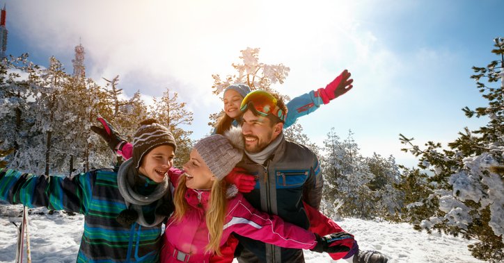A mom, dad, son, and daughter taking a break from downhill skiing to play in the snow on a sunny mountain slope surrounded by trees.