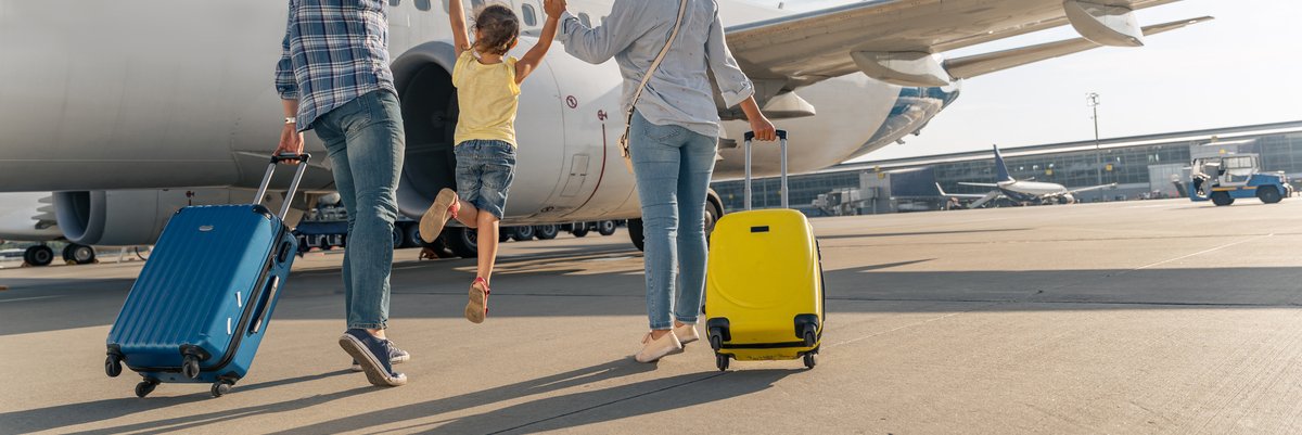 Two parents holding their jumping child's hands between them while rolling their suitcases toward an airplane on the tarmac.