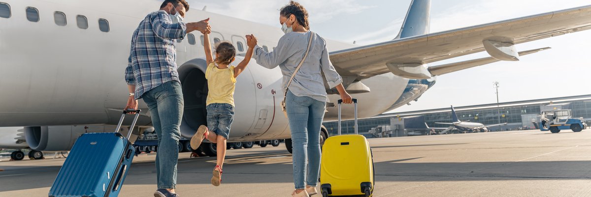Two parents holding their jumping child's hands between them while rolling their suitcases toward an airplane on the tarmac.