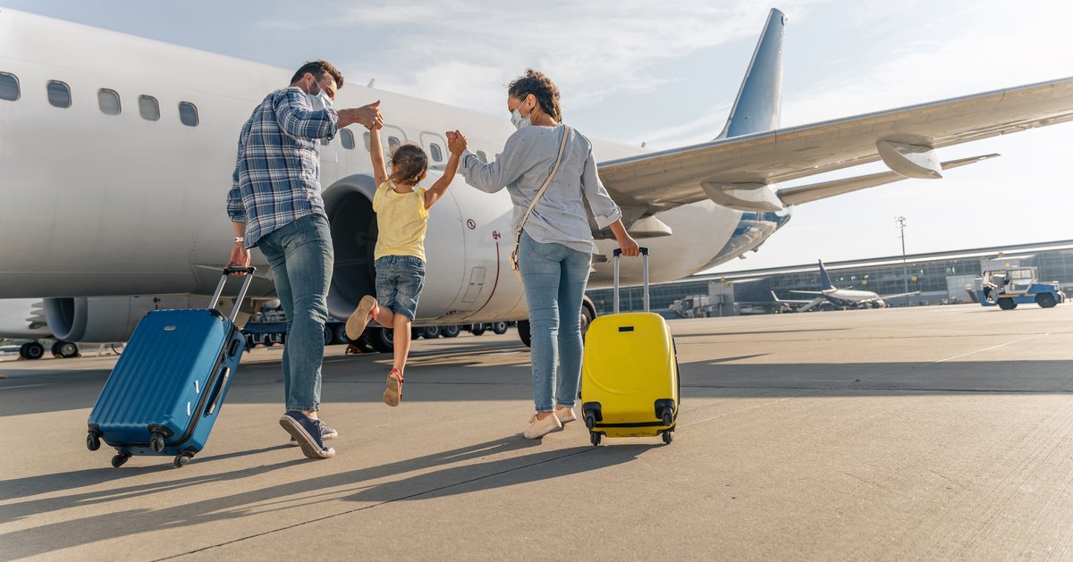 Two parents holding their jumping child's hands between them while rolling their suitcases toward an airplane on the tarmac.