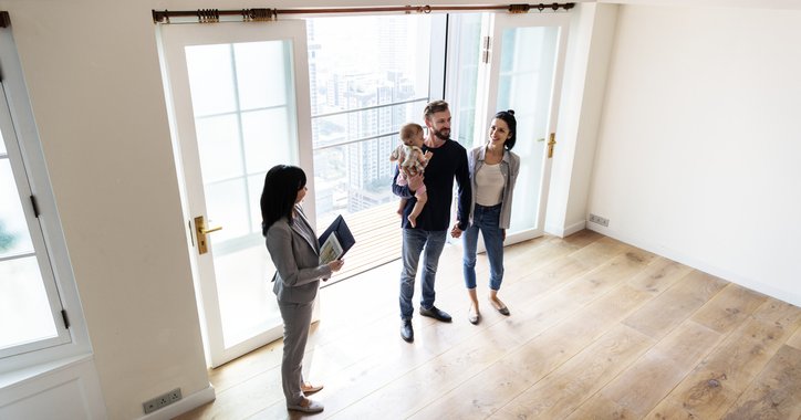 A man and woman with their young daughter touring an empty living room with a realtor.