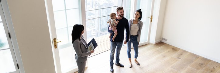 A man and woman with their young daughter touring an empty living room with a realtor.
