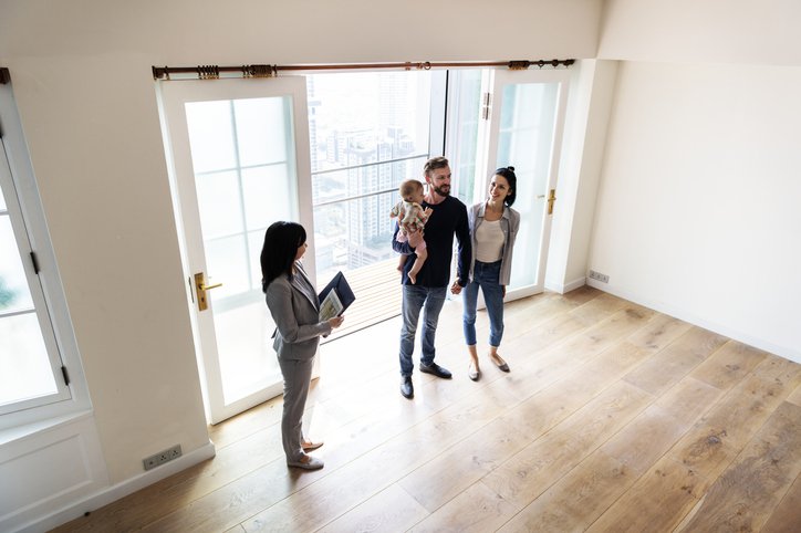 A man and woman with their young daughter touring an empty living room with a realtor.