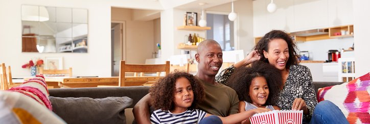 A mom, dad, and two daughters sitting on their living room couch and sharing popcorn while watching a movie.