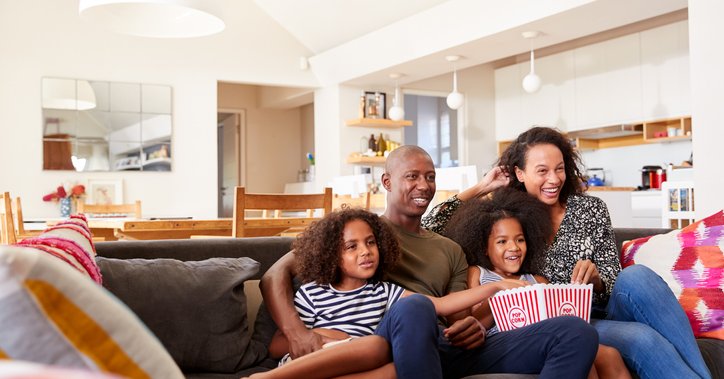 A mom, dad, and two daughters sitting on their living room couch and sharing popcorn while watching a movie.