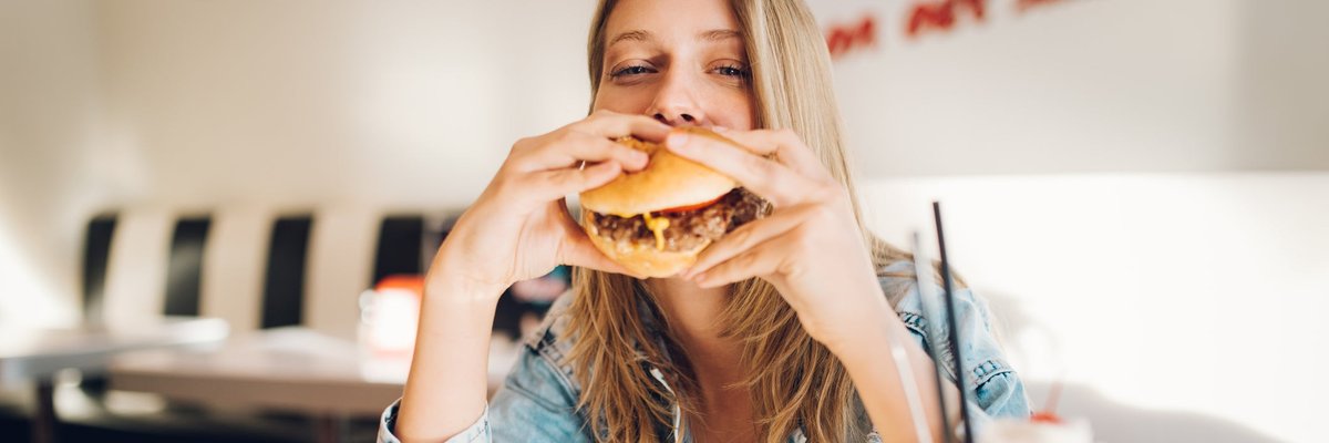 A person eating a hamburger with a plate of fries and a milkshake on the table in a fast food restaurant.