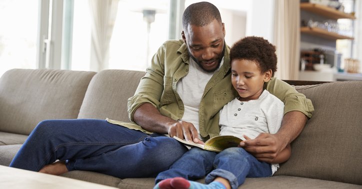 A father sitting on a couch with his arm around his son while they read a book.