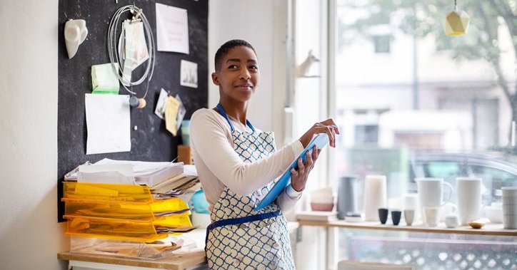 A woman wearing an apron and standing in her small pottery shop holding a clipboard.
