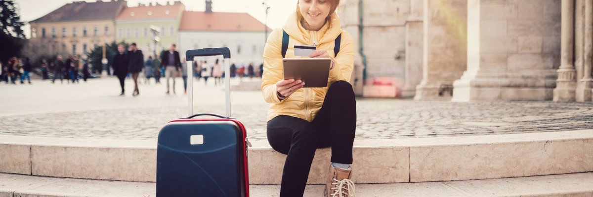 A woman sitting in a sunny plaza next to her suitcase while she looks something up on her tablet and holds her credit card.