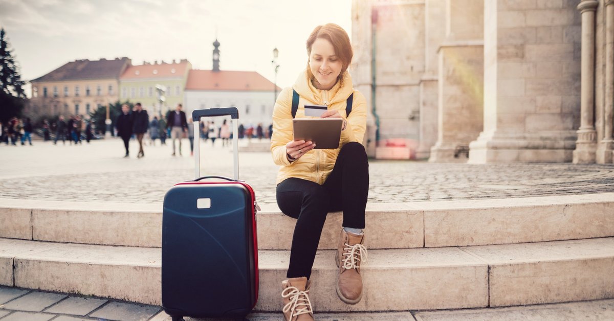 A woman sitting in a sunny plaza next to her suitcase while she looks something up on her tablet and holds her credit card.