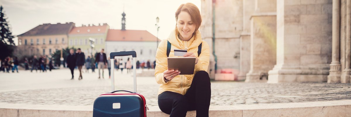 A woman sitting in a sunny plaza next to her suitcase while she looks something up on her tablet and holds her credit card.